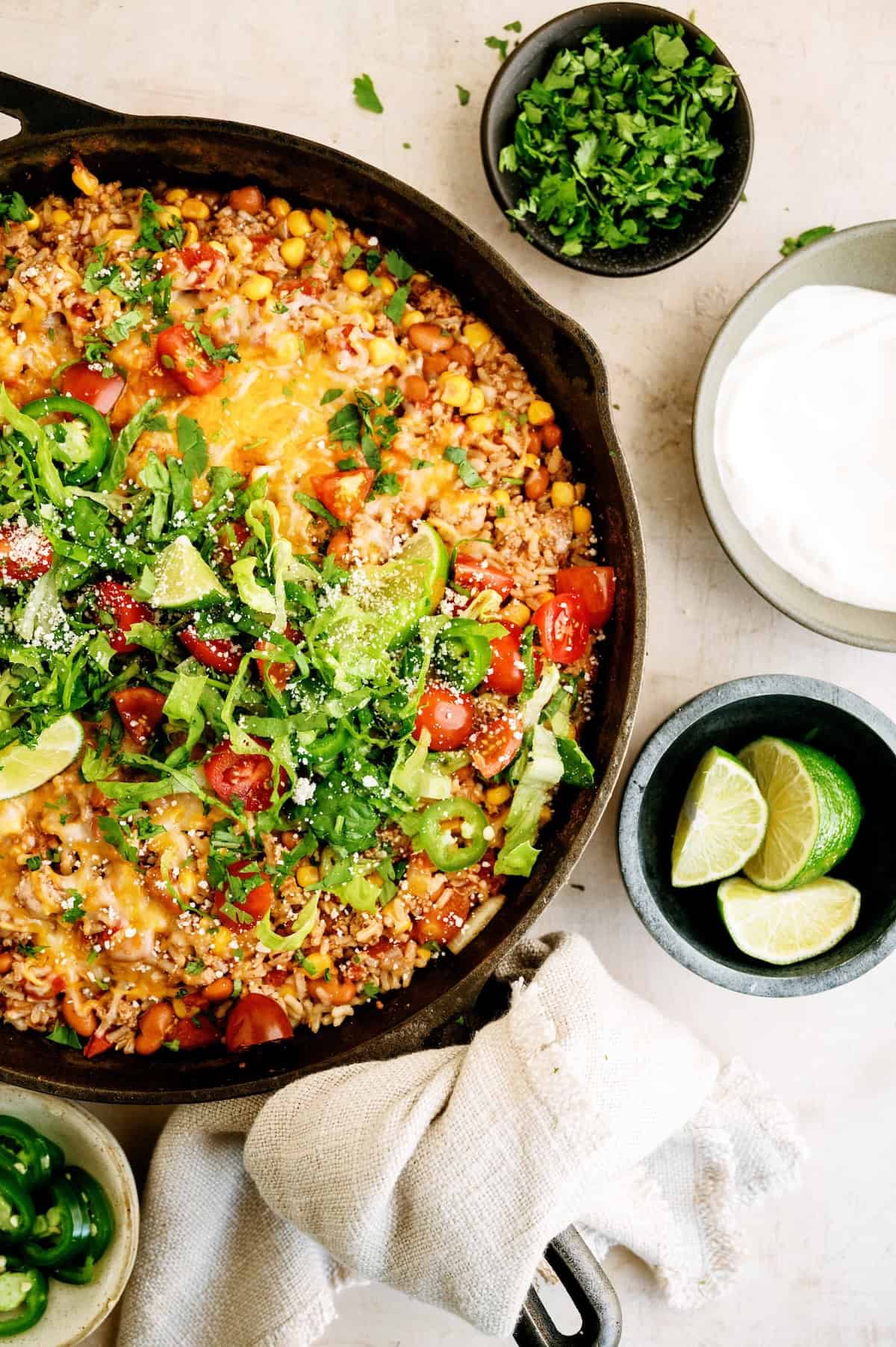 A cast iron skillet filled with baked Mexican-style rice topped with cheese, tomatoes, greens, and lime wedges, surrounded by bowls of fresh cilantro, lime slices, and sour cream.