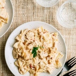 A plate of bowtie pasta in a creamy sauce garnished with parsley, served on a woven placemat with black cutlery and two empty glasses.