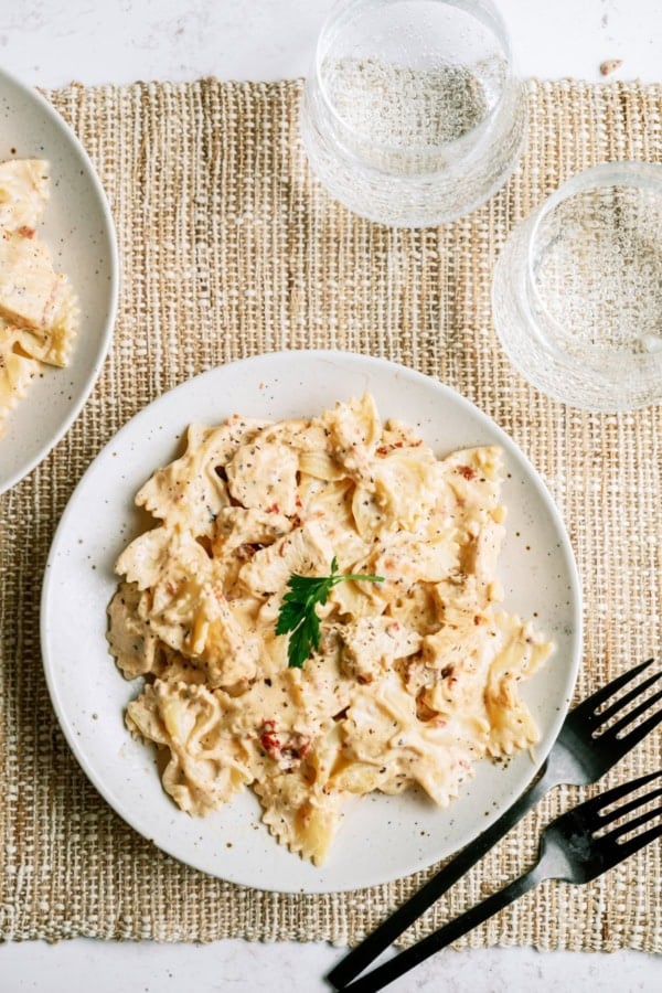 A plate of bowtie pasta in a creamy sauce garnished with parsley, served on a woven placemat with black cutlery and two empty glasses.