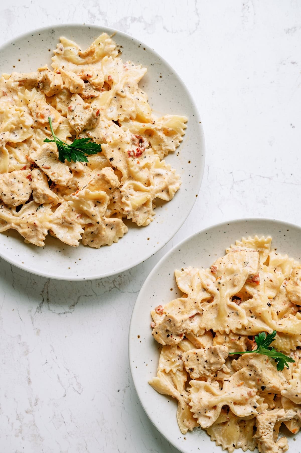 Two plates of creamy bowtie pasta with pieces of chicken, garnished with a sprig of parsley and sprinkled with black pepper, on a white surface.