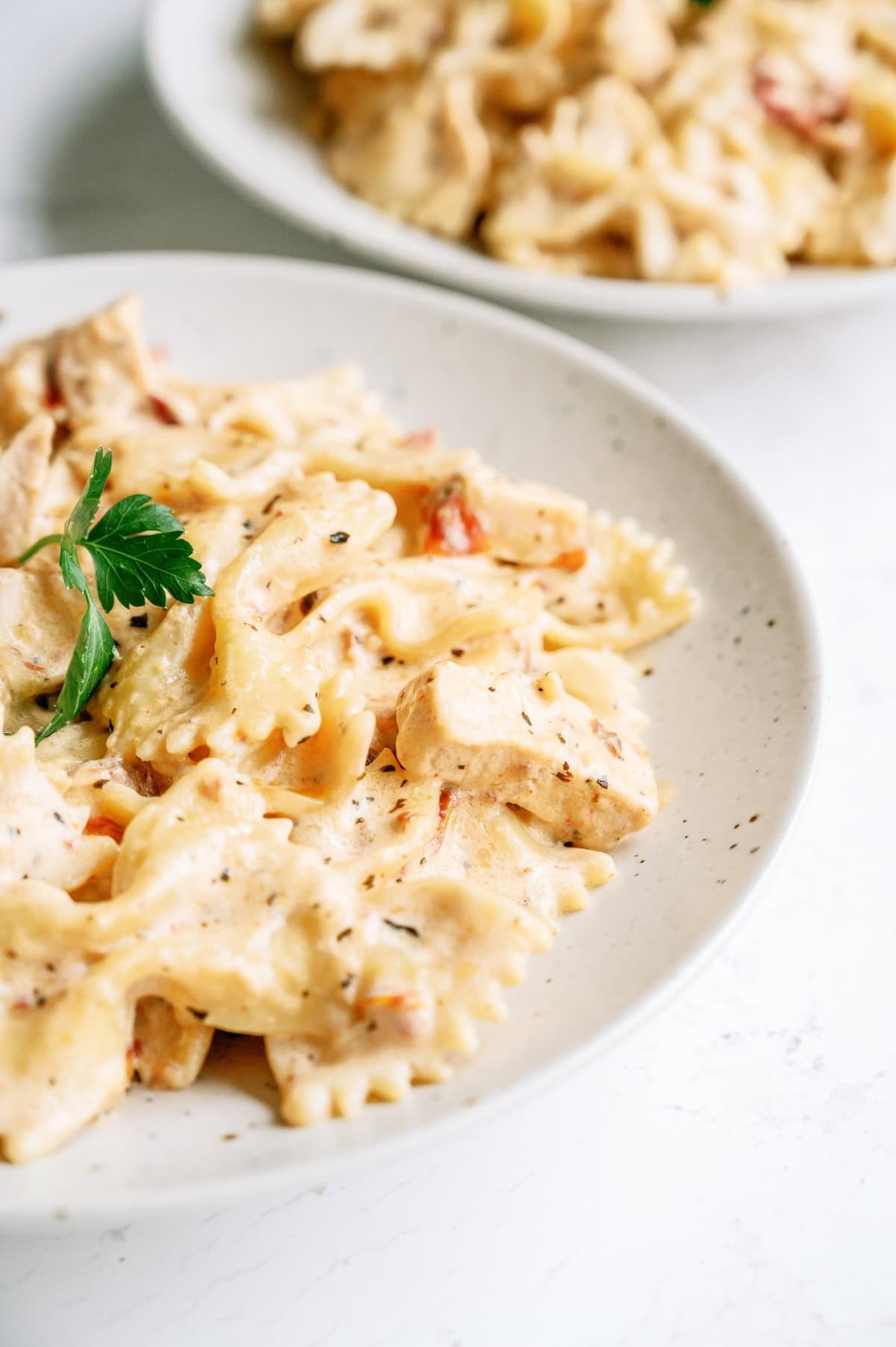 A white plate of creamy bow-tie pasta garnished with a sprig of parsley, with another similar plate in the background.