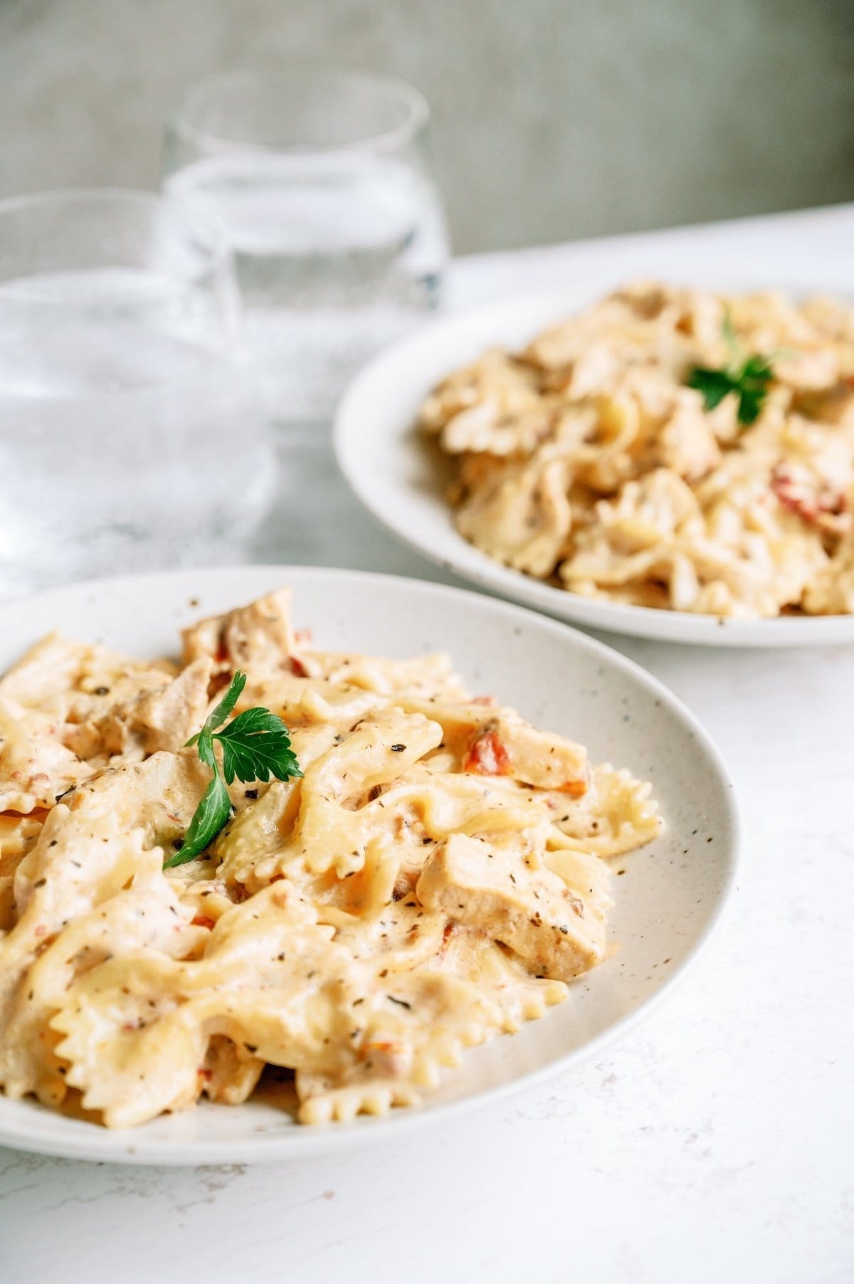 Two bowls of creamy bow-tie pasta with chicken and herbs, garnished with parsley, are placed on a white surface next to glasses of water.