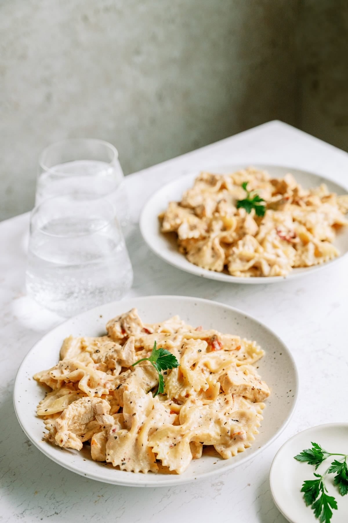 Two plates of bowtie pasta with creamy sauce and chicken pieces, garnished with parsley, are set on a white table beside two glasses of water.