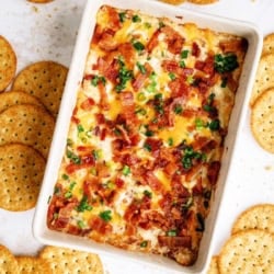 Rectangular baking dish filled with a baked cheese dip topped with bacon and green onions, surrounded by round crackers on a white surface.