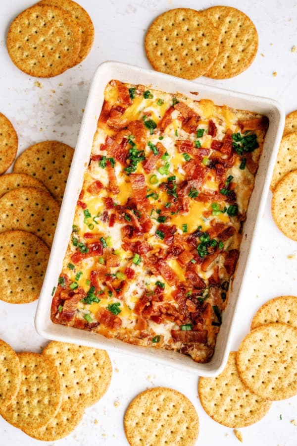 Rectangular baking dish filled with a baked cheese dip topped with bacon and green onions, surrounded by round crackers on a white surface.