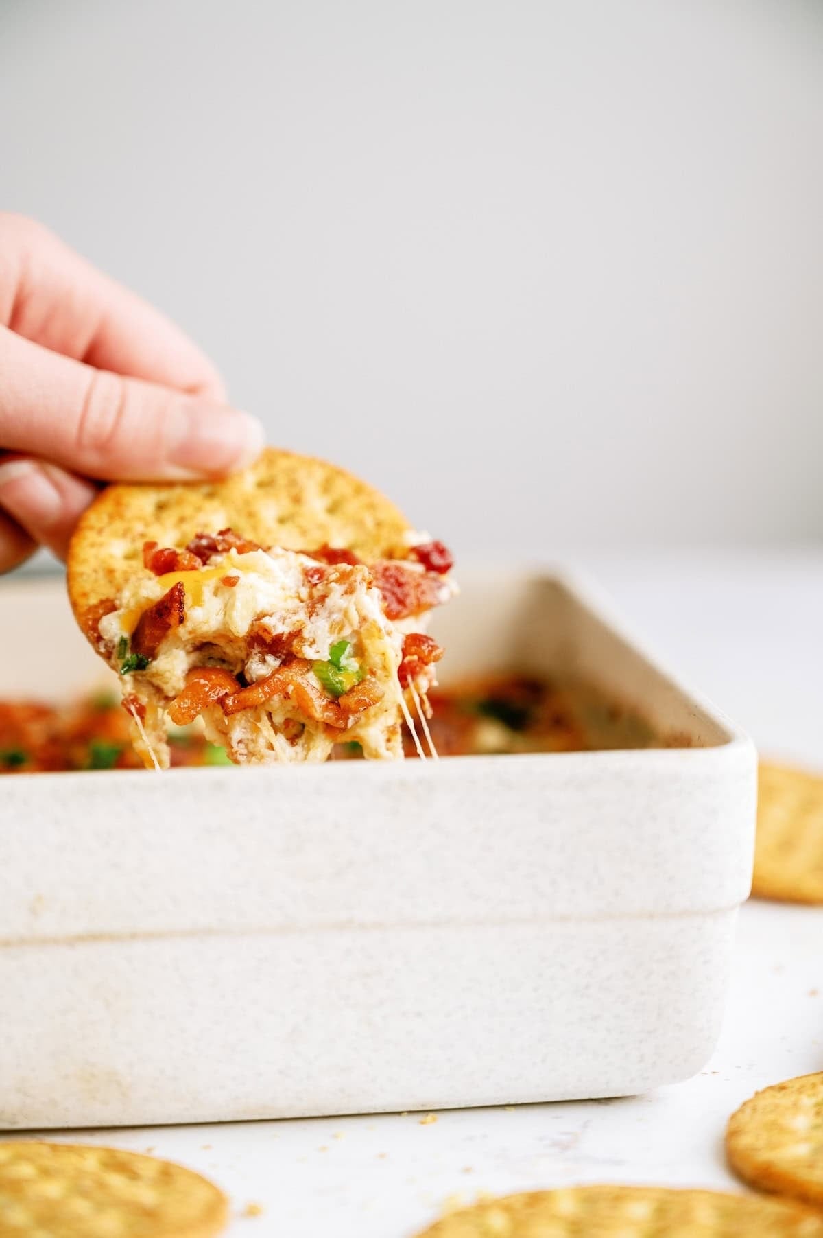 A hand dips a round cracker into a cheesy baked dip in a white square dish, with more crackers scattered on the table.