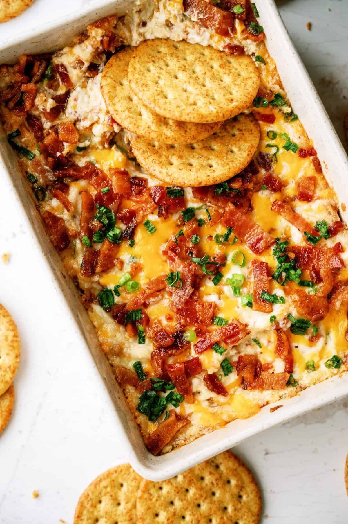 Rectangular dish of cheesy dip topped with bacon and chopped green onions, with round crackers partially dipped and scattered around.