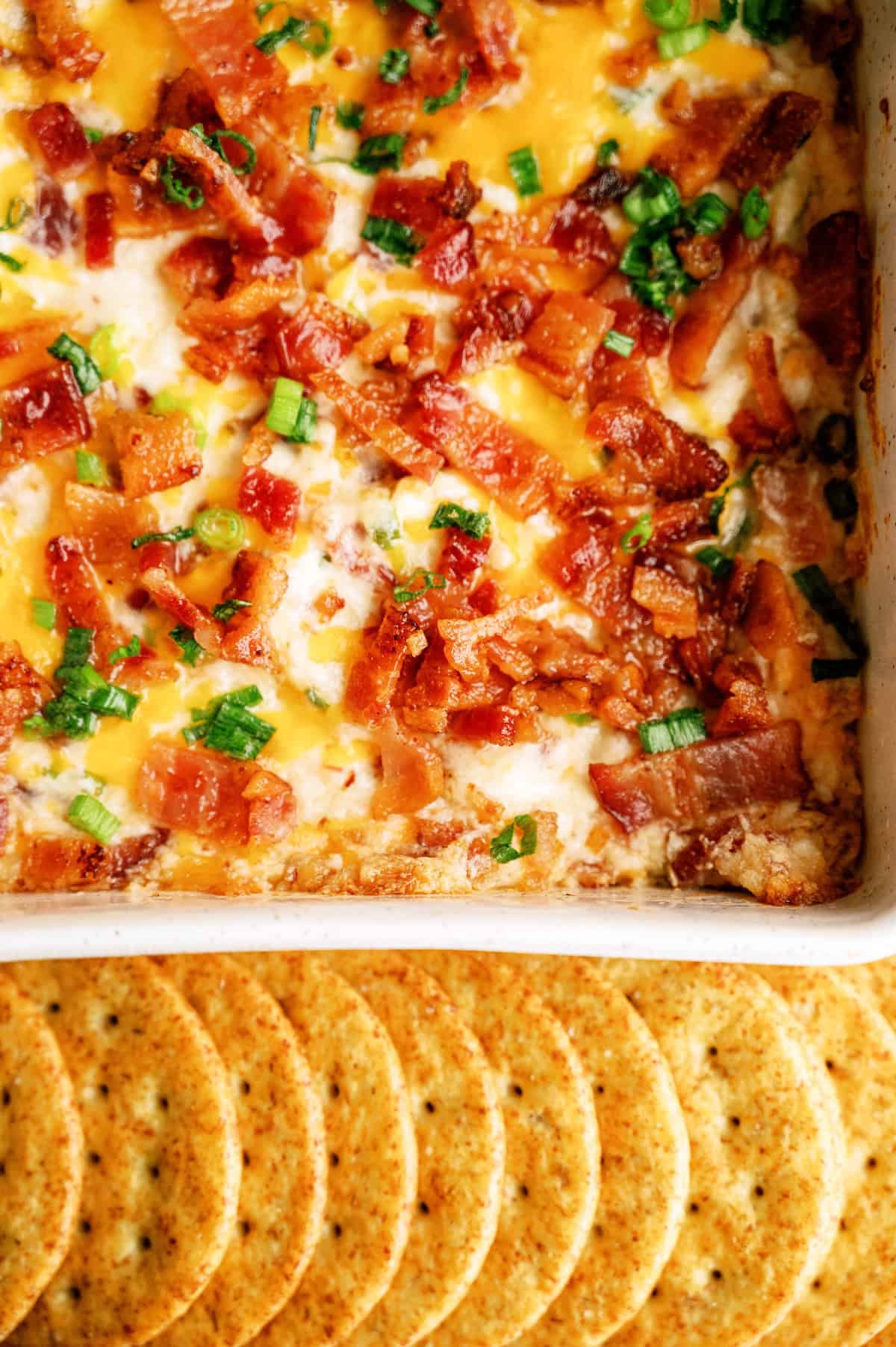 Baked dip topped with melted cheese, chopped bacon, and green onions in a dish, with a row of round crackers arranged below.