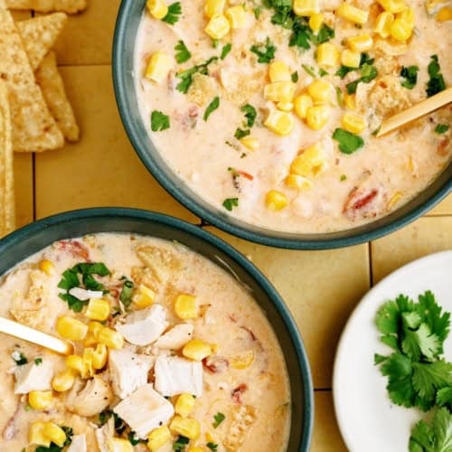 Two bowls of creamy soup topped with corn, chopped chicken, and cilantro, next to tortilla chips and a small plate of fresh cilantro.