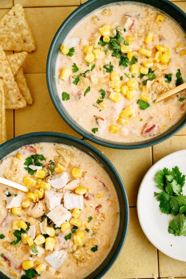 Two bowls of creamy soup topped with corn, chopped chicken, and cilantro, next to tortilla chips and a small plate of fresh cilantro.