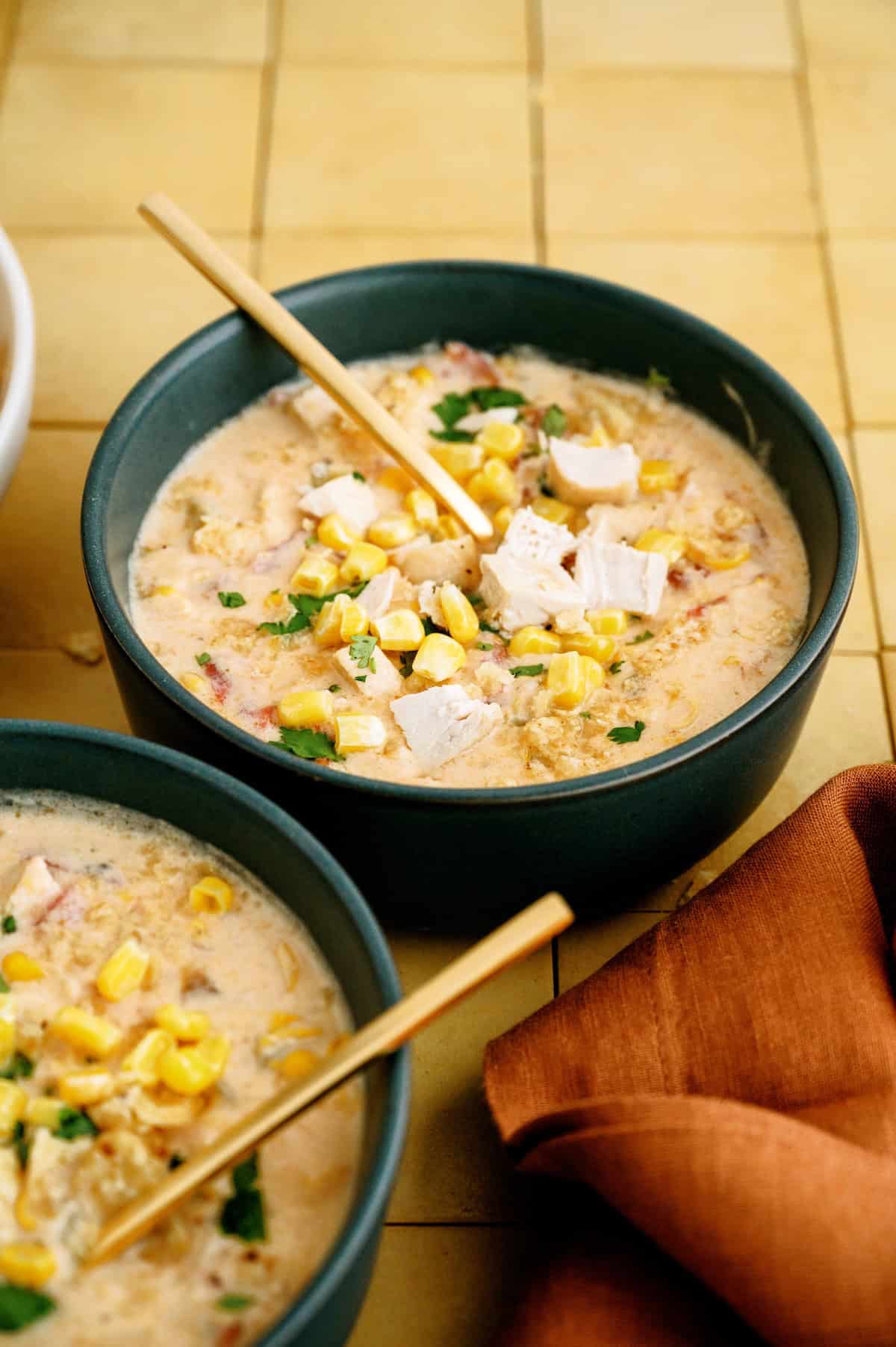Two bowls of creamy soup topped with corn, chopped chicken, and herbs, each with a wooden spoon, set on a yellow tiled surface beside a folded orange napkin.
