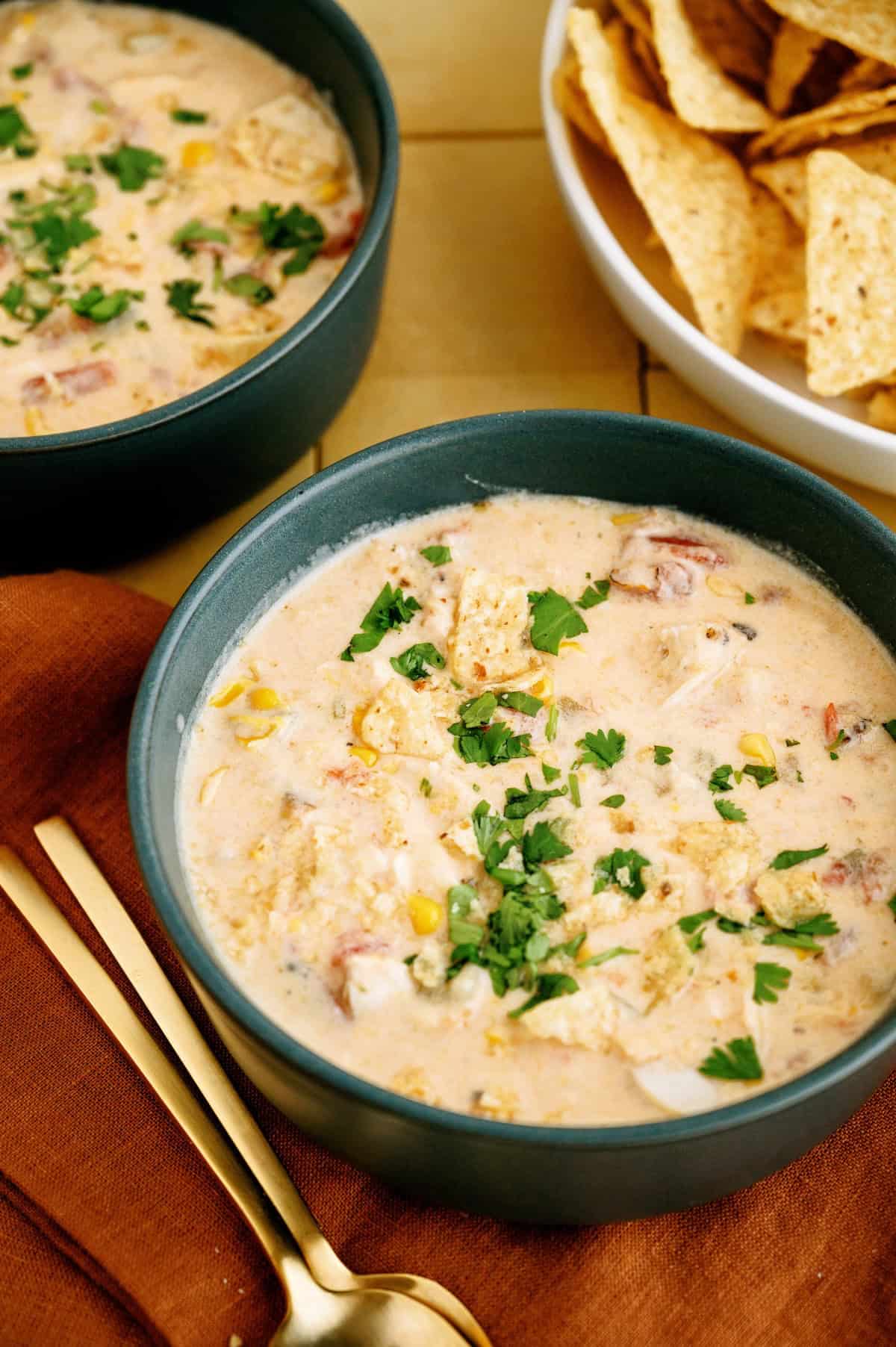 Two bowls of creamy soup garnished with chopped herbs, with a bowl of tortilla chips on the side and gold utensils on a brown napkin.