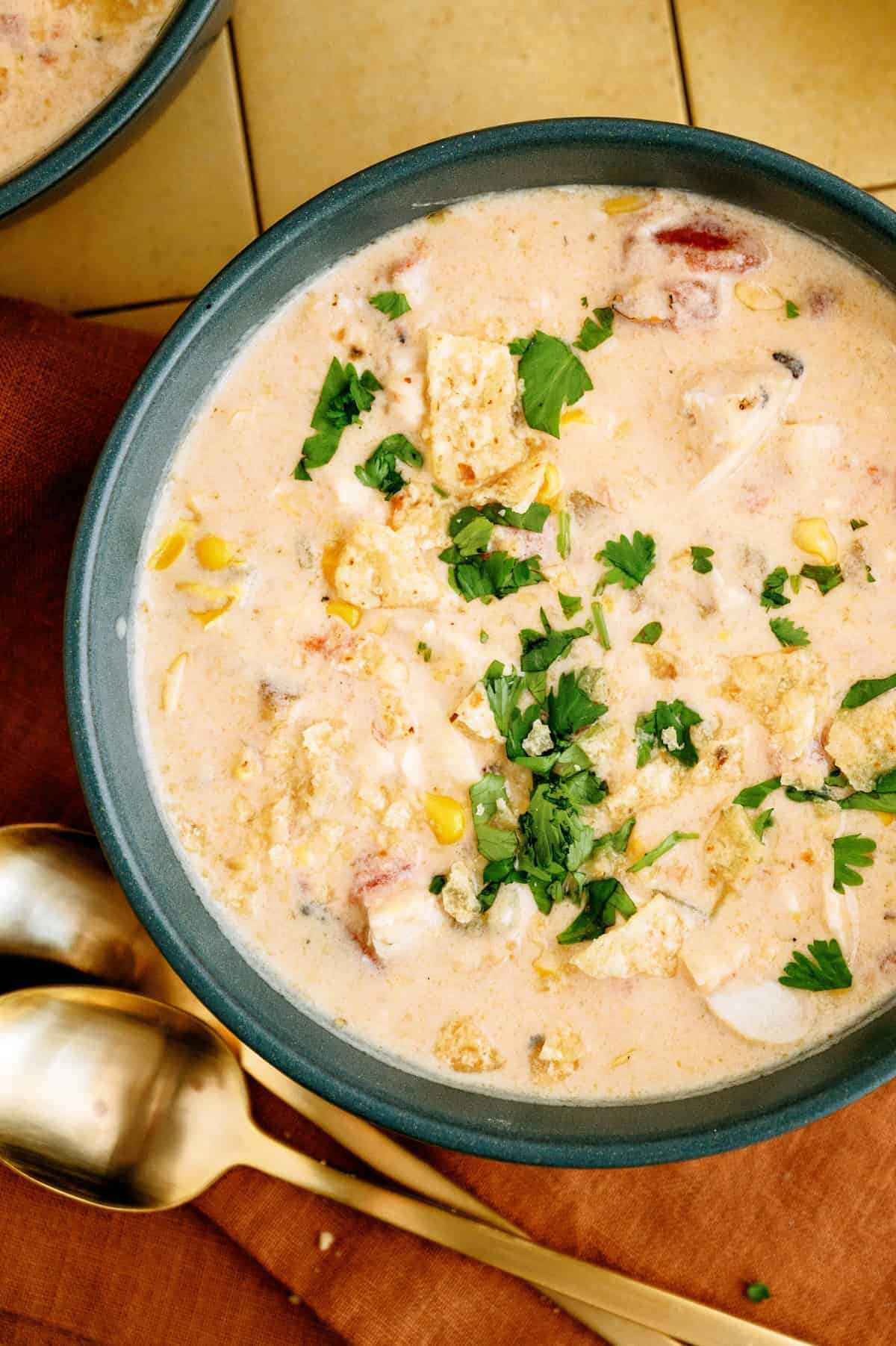 A bowl of creamy soup containing corn, chicken, herbs, and crushed tortilla chips sits on a table next to two gold spoons.