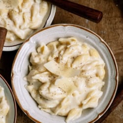 A bowl of creamy pasta topped with shaved cheese and black pepper, with a fork and spoon nearby on a wooden surface.