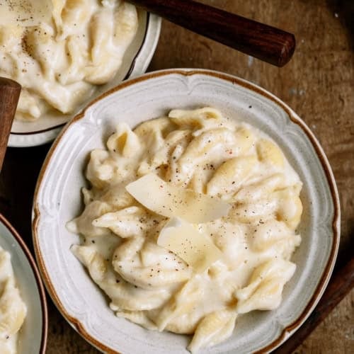 A bowl of creamy pasta topped with shaved cheese and black pepper, with a fork and spoon nearby on a wooden surface.