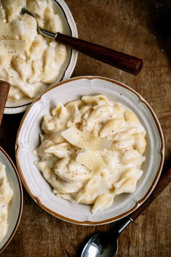 A bowl of creamy pasta topped with shaved cheese and black pepper, with a fork and spoon nearby on a wooden surface.