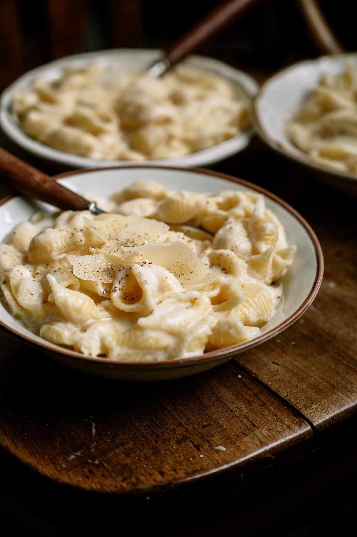 A bowl of shell pasta in creamy white sauce, topped with black pepper and cheese shavings, sits on a wooden table with two similar bowls in the background.