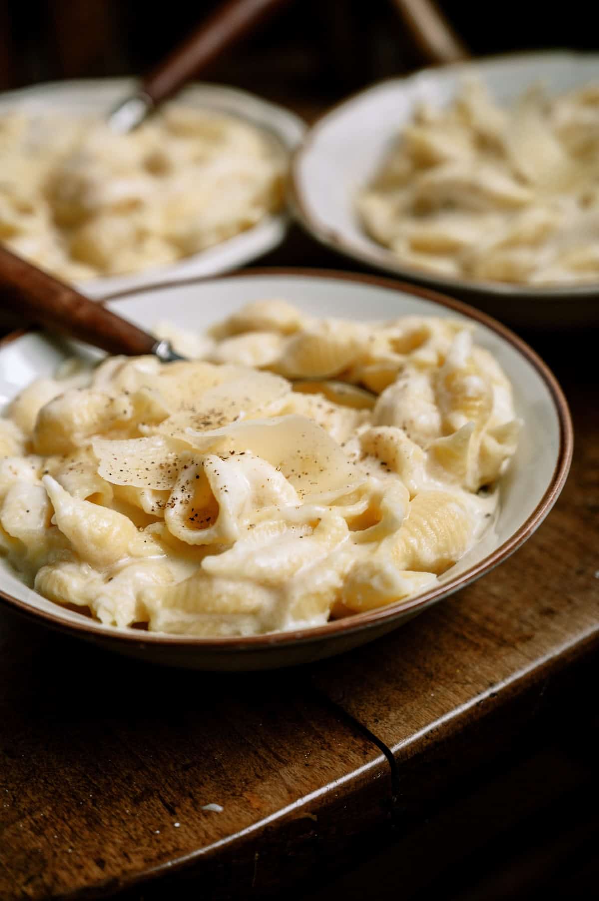 Three bowls of shell pasta in a creamy white sauce, topped with black pepper and cheese, on a wooden table.