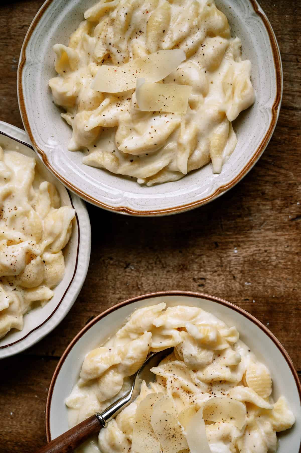 Three bowls of creamy pasta shells topped with shaved cheese and black pepper sit on a wooden surface.