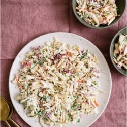 A large serving of Apple Slaw on a plate with two small bowls of Apple Slaw on the side.