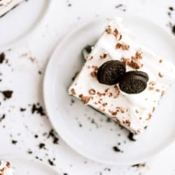 An Oreo ice cream cake topped with whipped cream, chocolate shavings, and two mini chocolate sandwich cookies on a white plate, with cookie crumbs scattered around.