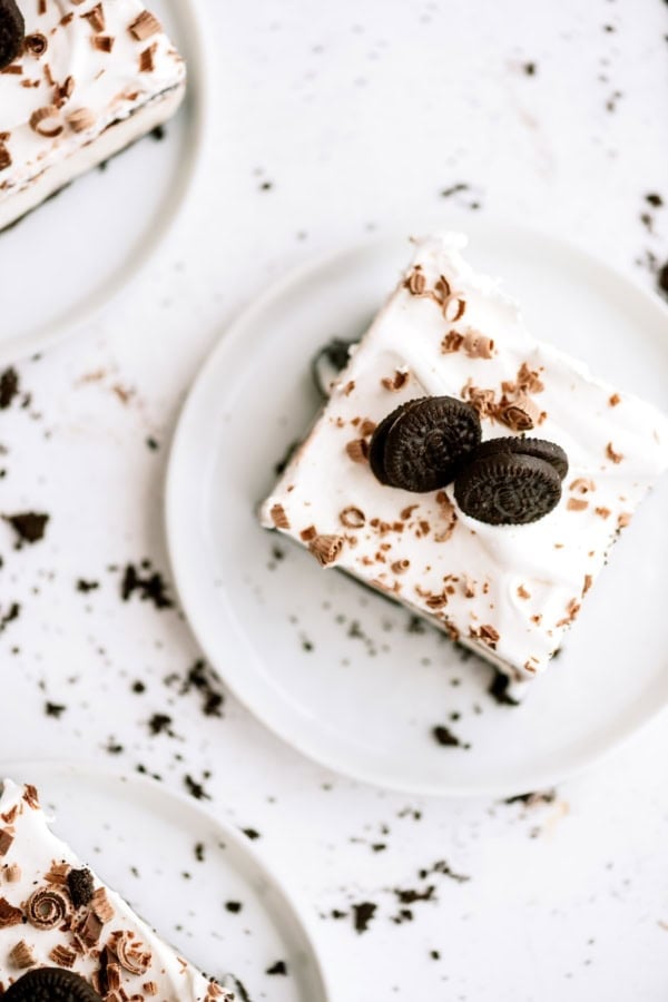 An Oreo ice cream cake topped with whipped cream, chocolate shavings, and two mini chocolate sandwich cookies on a white plate, with cookie crumbs scattered around.