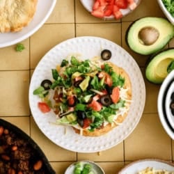A plate with a tostada topped with lettuce, tomato, olives, cheese, and beans is surrounded by bowls of avocado, chopped tomato, ground meat, green onions, lettuce, and shredded cheese.