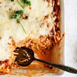 Baked pasta casserole topped with melted cheese and garnished with parsley, with a serving spoon and a portion removed from the dish.