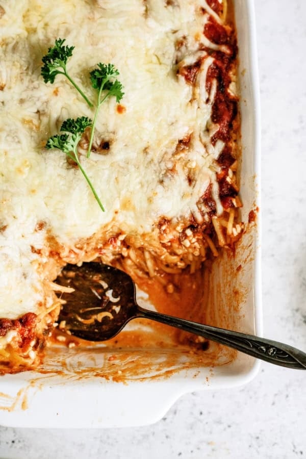 Baked pasta casserole topped with melted cheese and garnished with parsley, with a serving spoon and a portion removed from the dish.