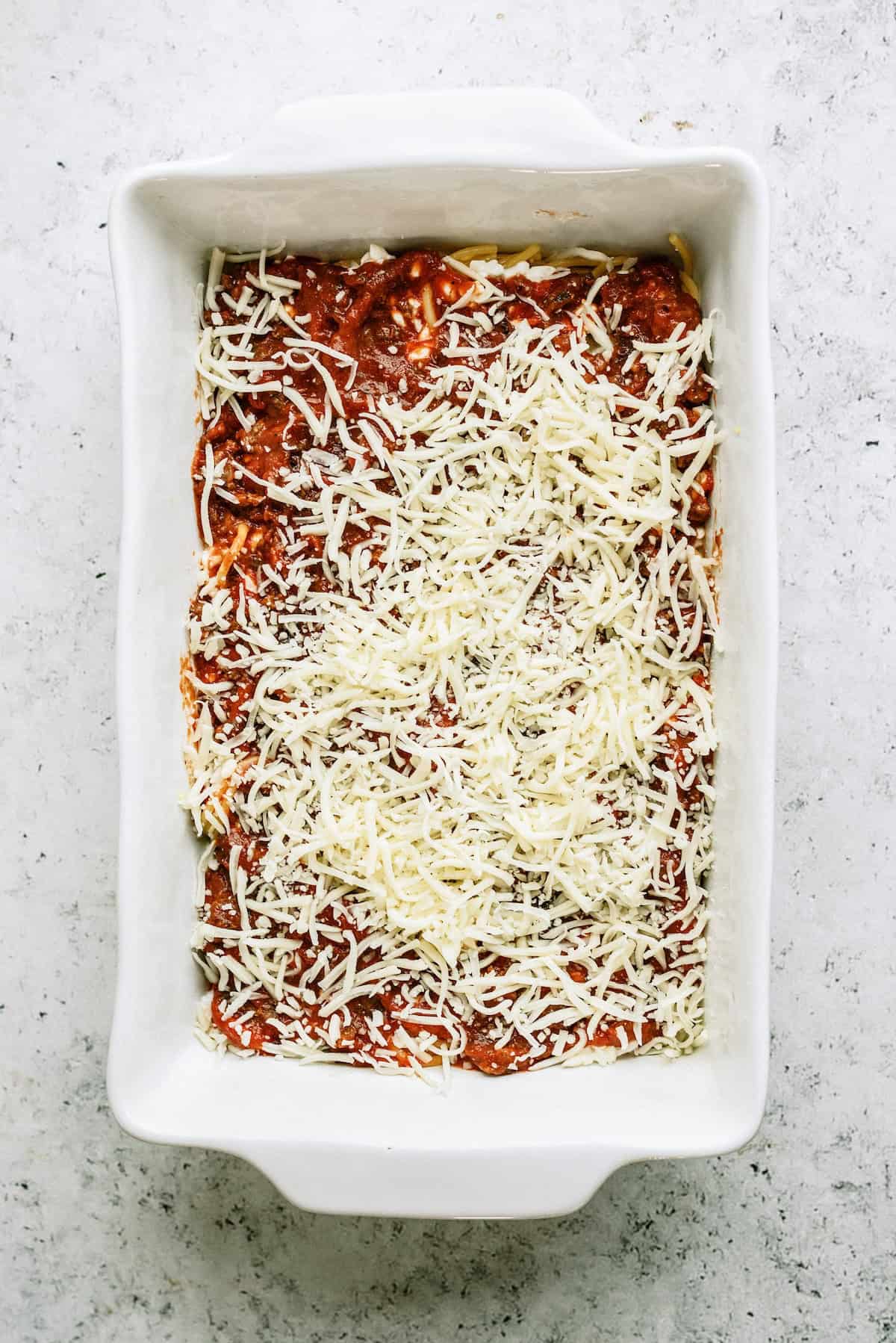 Rectangular white baking dish with a layer of tomato sauce and shredded cheese, ready to be baked on a light textured surface.