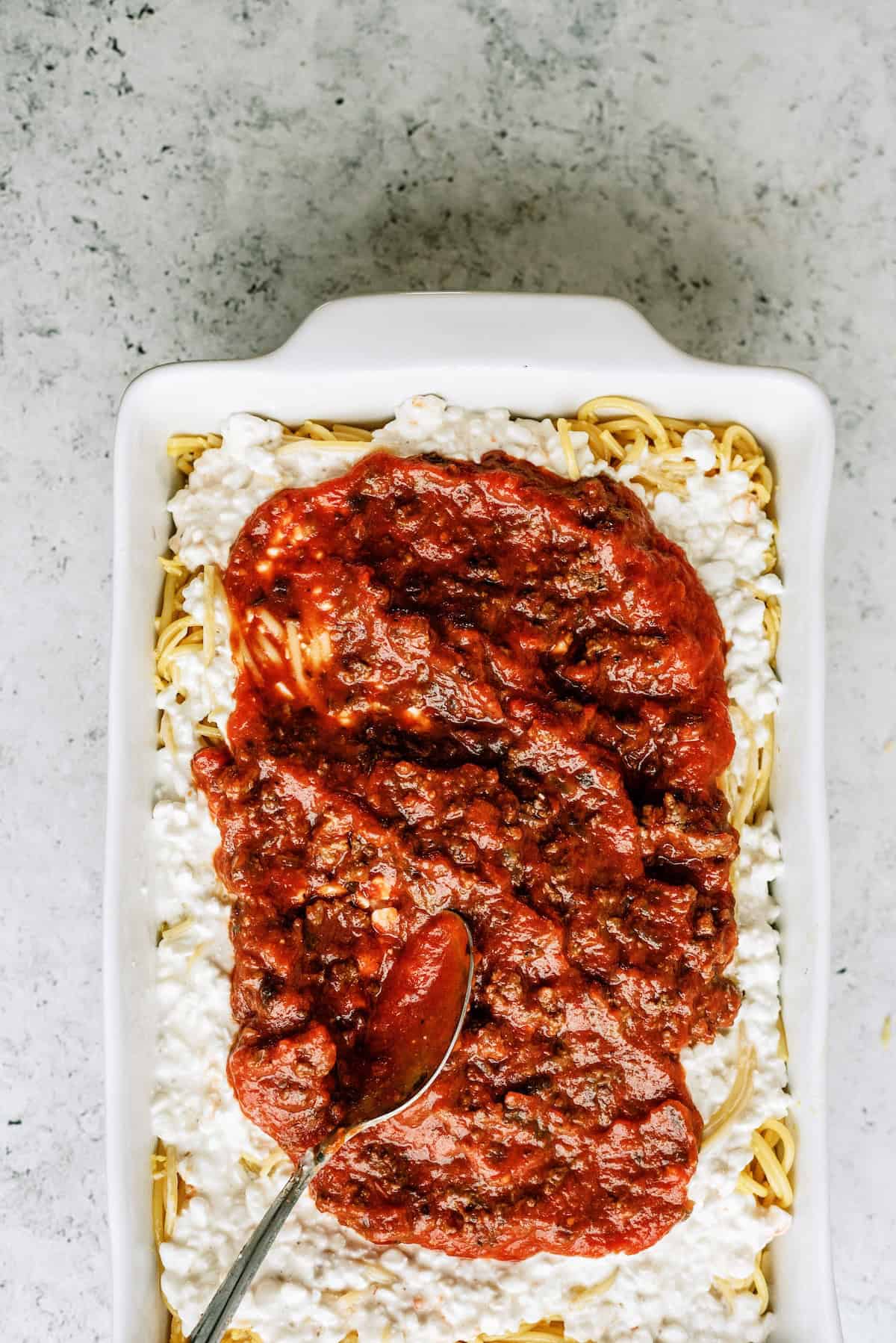 A white baking dish filled with cooked spaghetti, topped with cottage cheese and meat tomato sauce, with a spoon spreading the sauce.