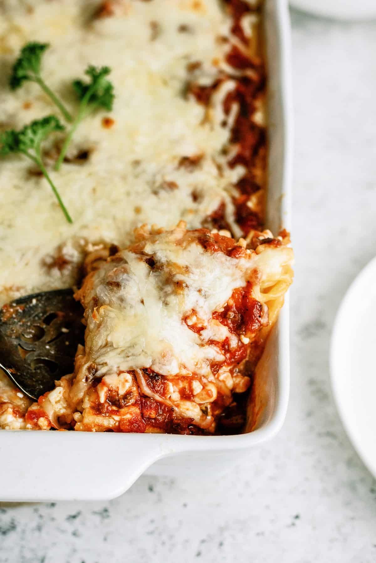 Close-up of a cheesy baked lasagna in a white dish, with a serving spatula lifting out a portion; topped with melted cheese and parsley garnish.