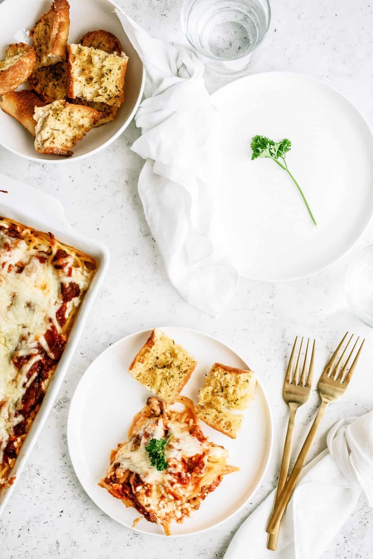 A plate with a serving of cheesy baked pasta and two slices of garlic bread, next to gold utensils, a white napkin, an empty plate with a parsley sprig, and a bowl of more garlic bread.