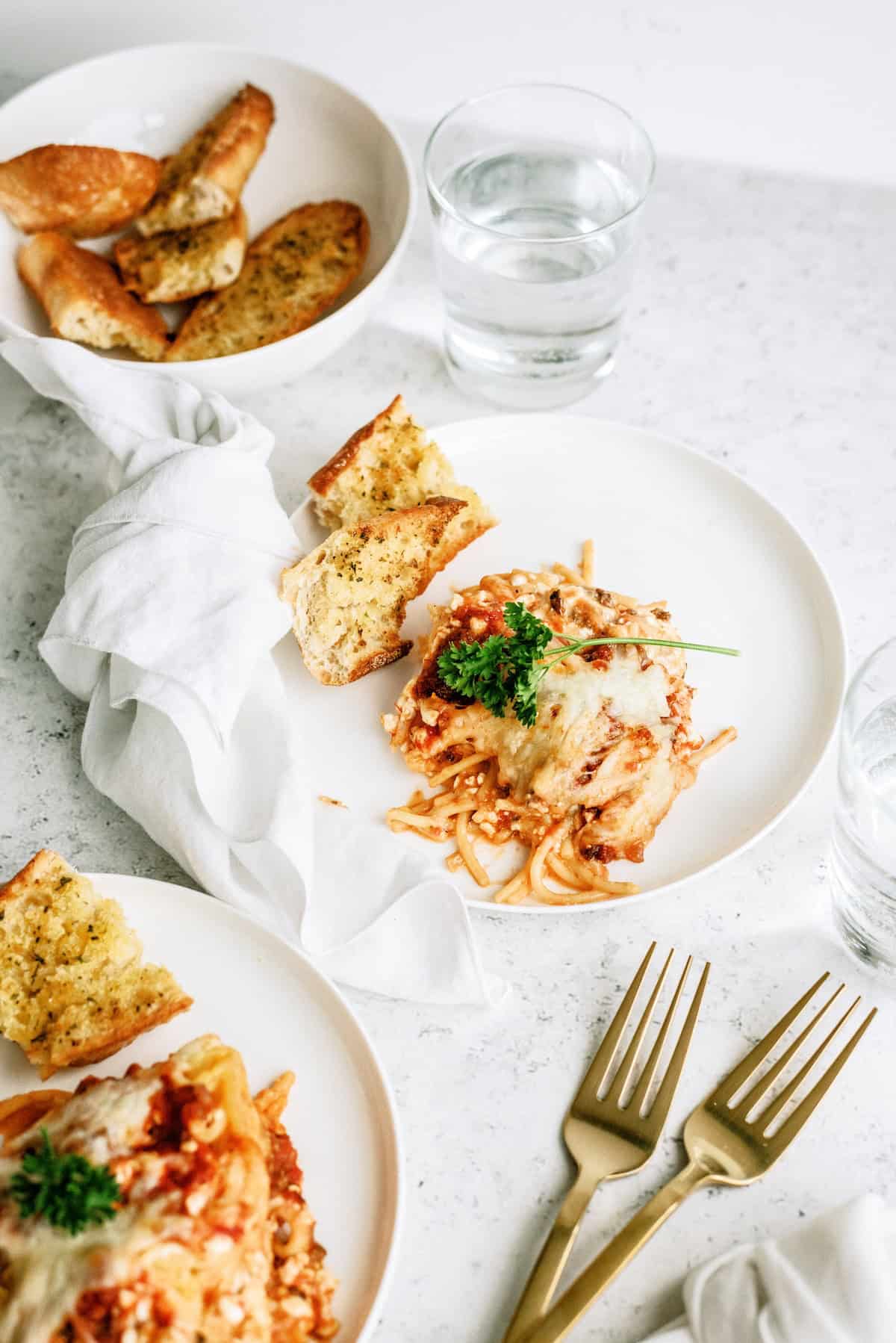 Two plates of baked pasta with tomato sauce and melted cheese, garnished with parsley, served with slices of garlic bread. Two glasses of water and gold forks are on the table.