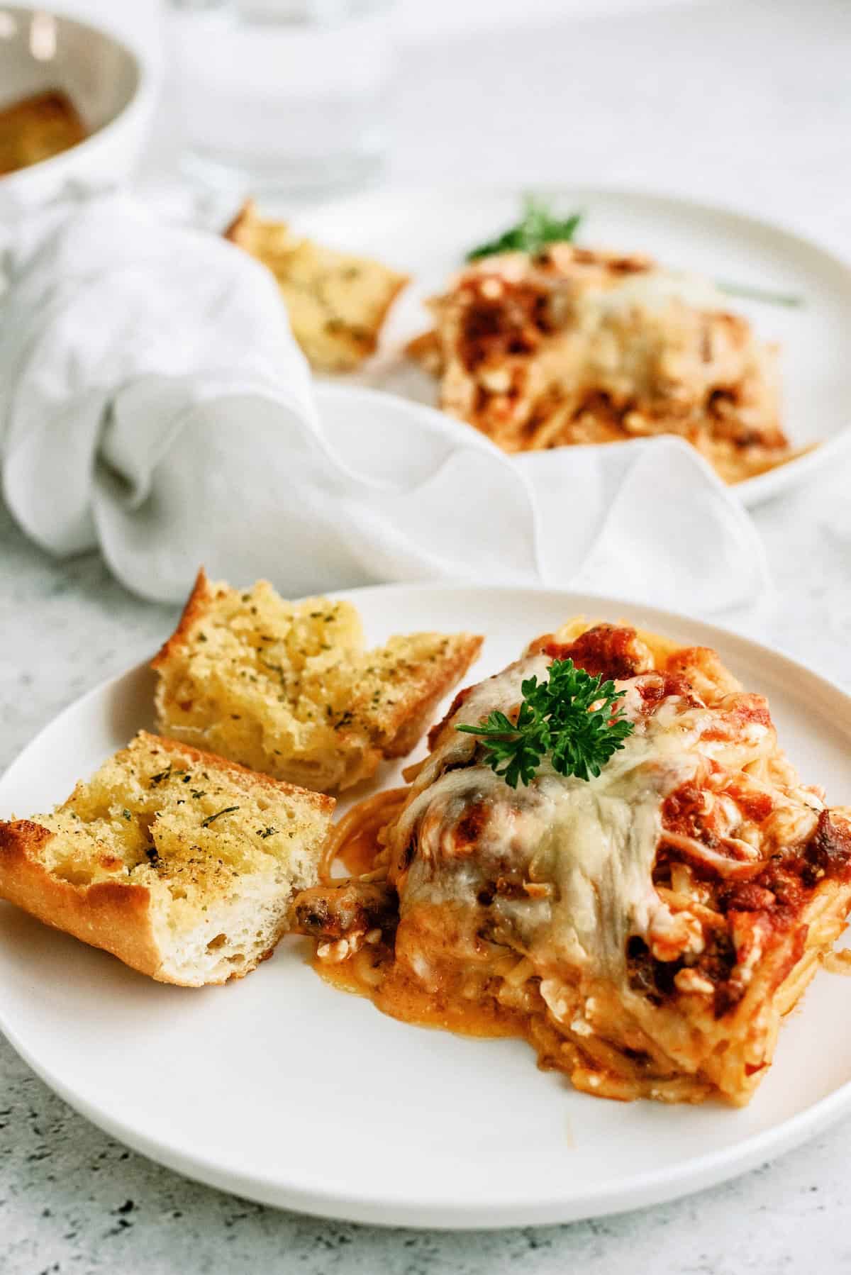 A plate with a serving of lasagna topped with parsley and two slices of garlic bread, with another plate and a white napkin in the background.