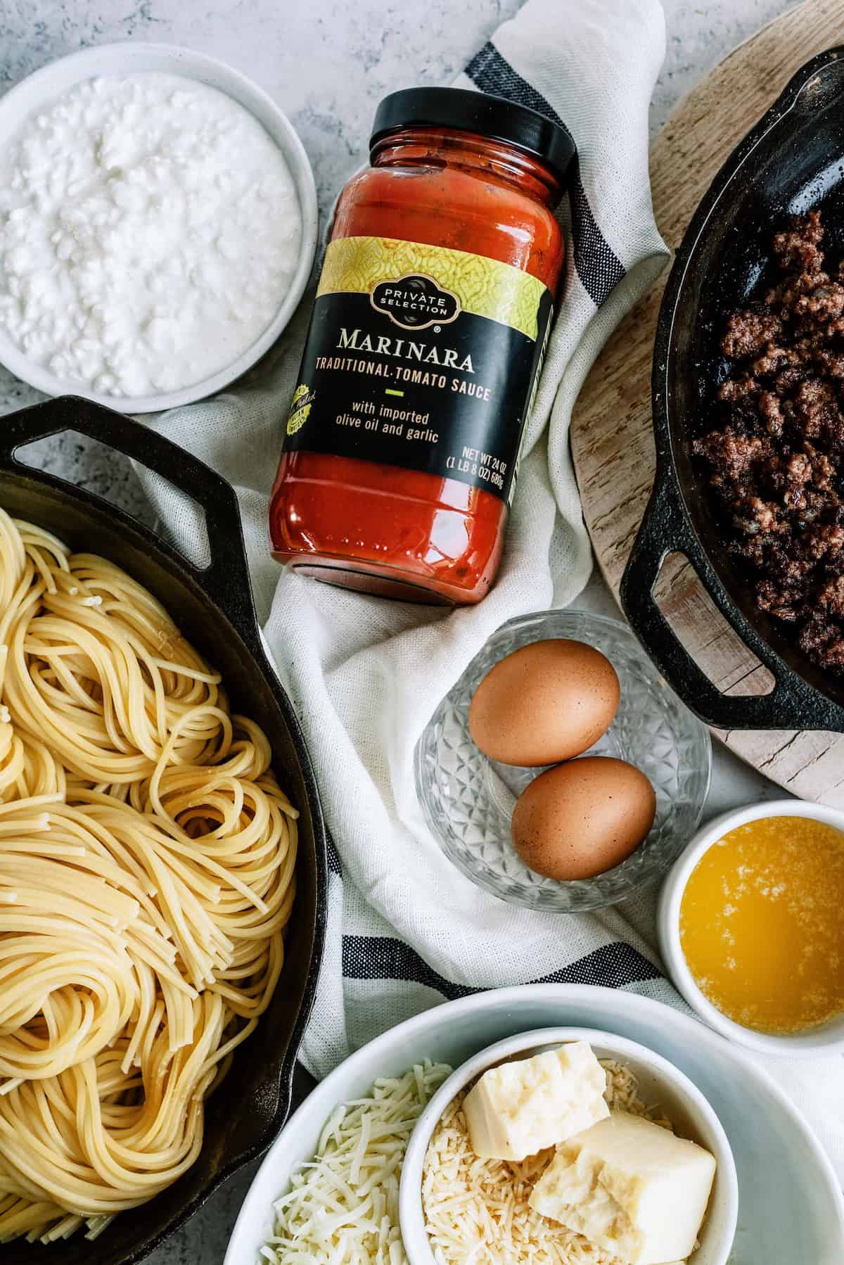 Spaghetti, marinara sauce, ground beef in a skillet, cottage cheese, eggs, melted butter, shredded cheese, and butter cubes arranged on a table.