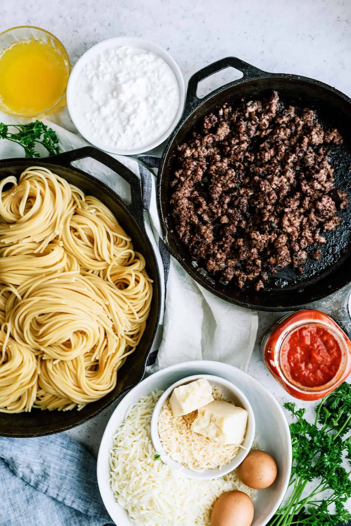 Cooked spaghetti, browned ground beef, tomato sauce, shredded cheese, eggs, cottage cheese, butter, and parsley arranged on a table, viewed from above.