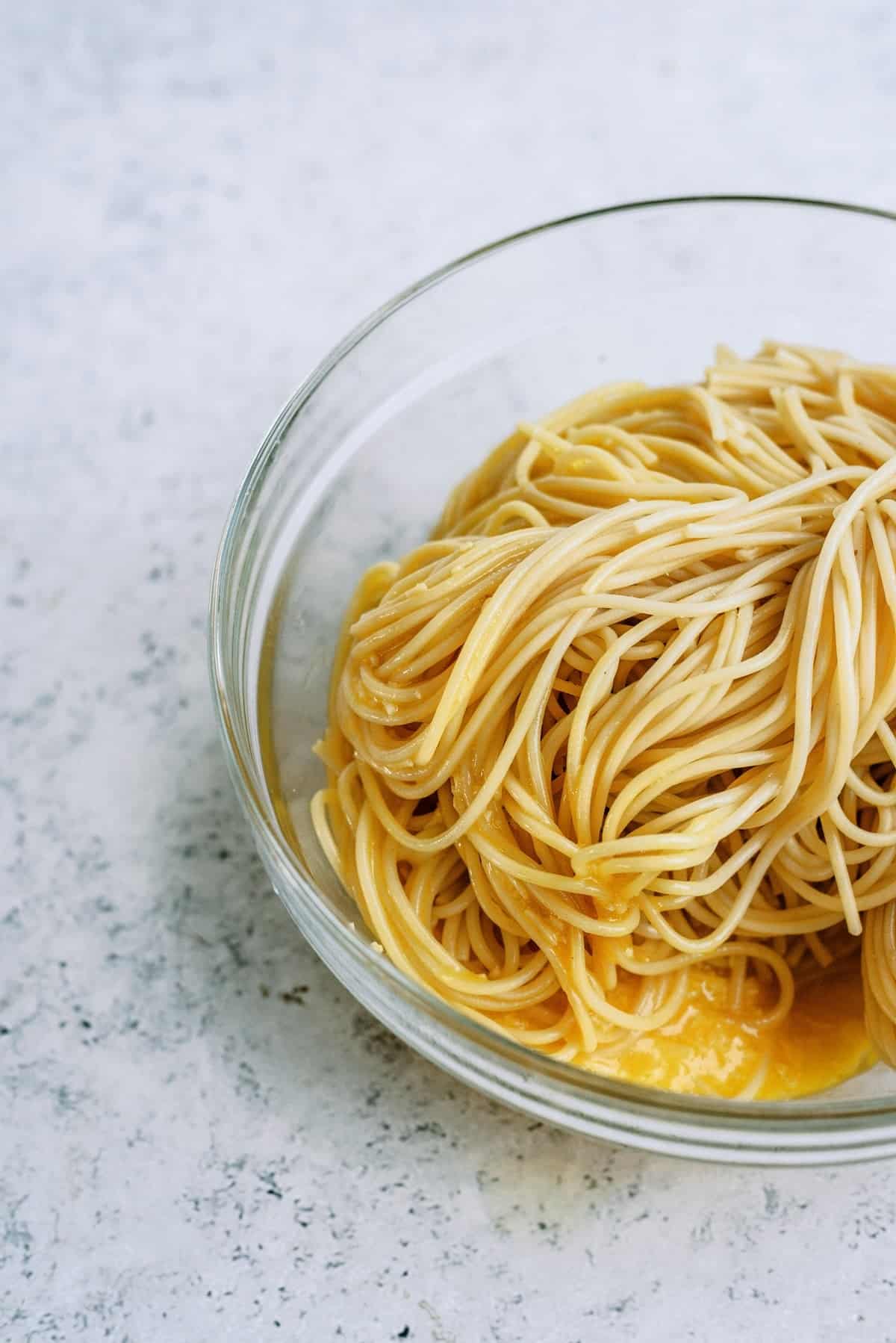 A clear glass bowl filled with plain cooked spaghetti noodles coated in a yellow sauce, placed on a light textured surface.