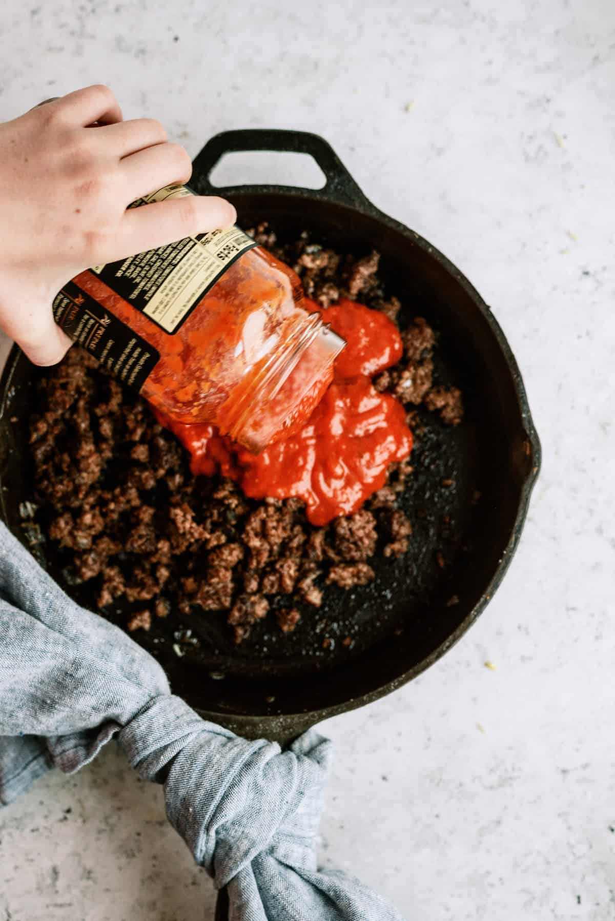 A hand pours tomato sauce from a jar into a cast iron skillet filled with cooked ground beef.