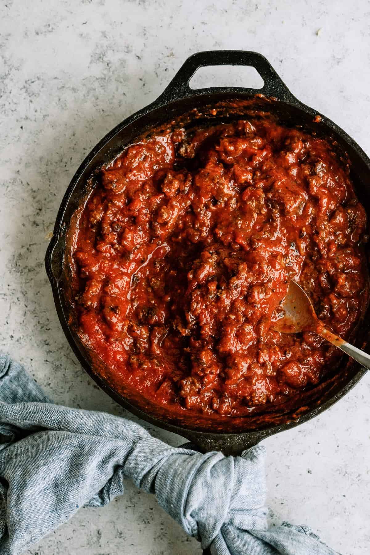 A cast iron skillet filled with chunky tomato meat sauce sits on a light countertop with a wooden spoon and a gray cloth nearby.