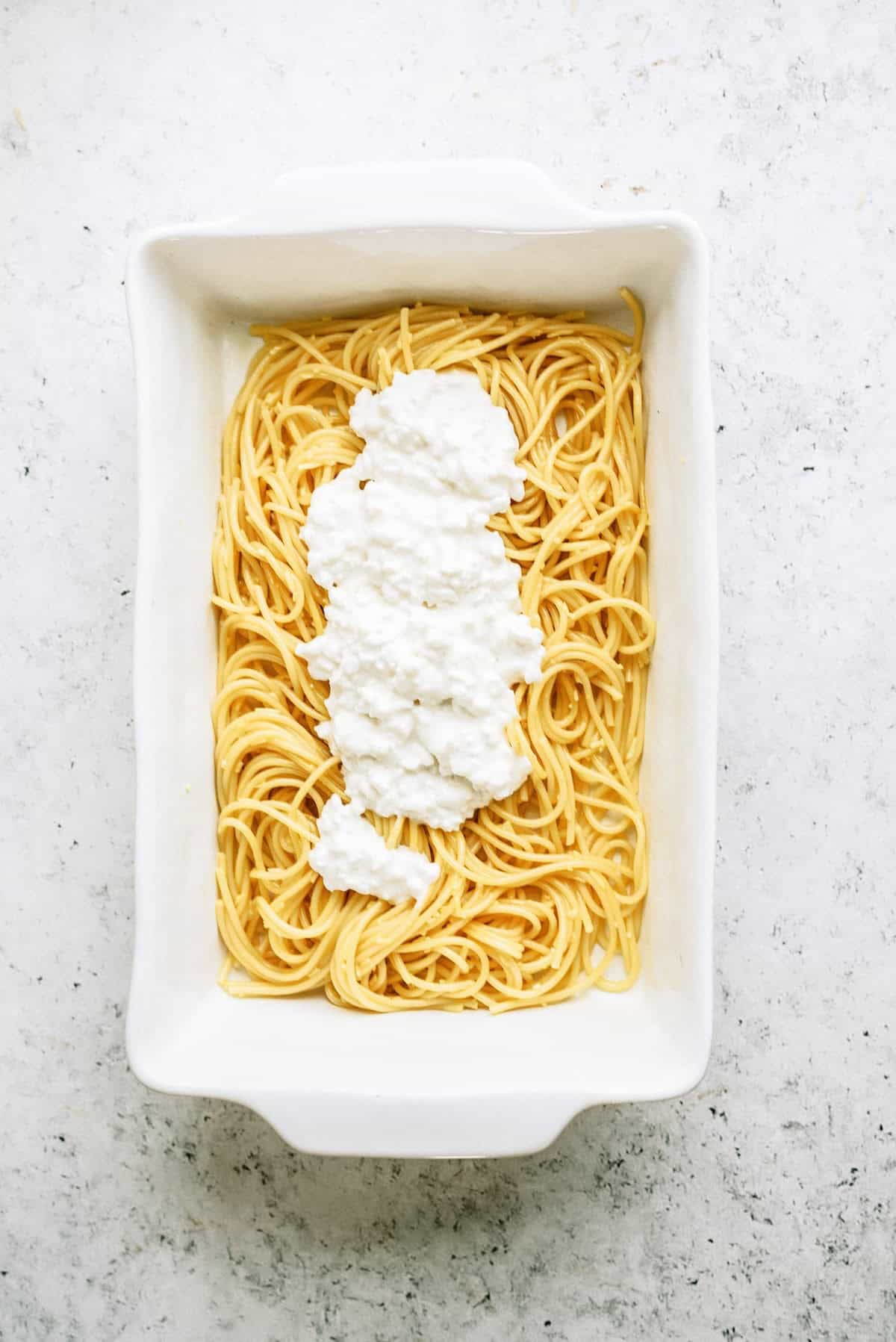 A white baking dish with cooked spaghetti topped with a layer of cottage cheese on a light-colored surface.
