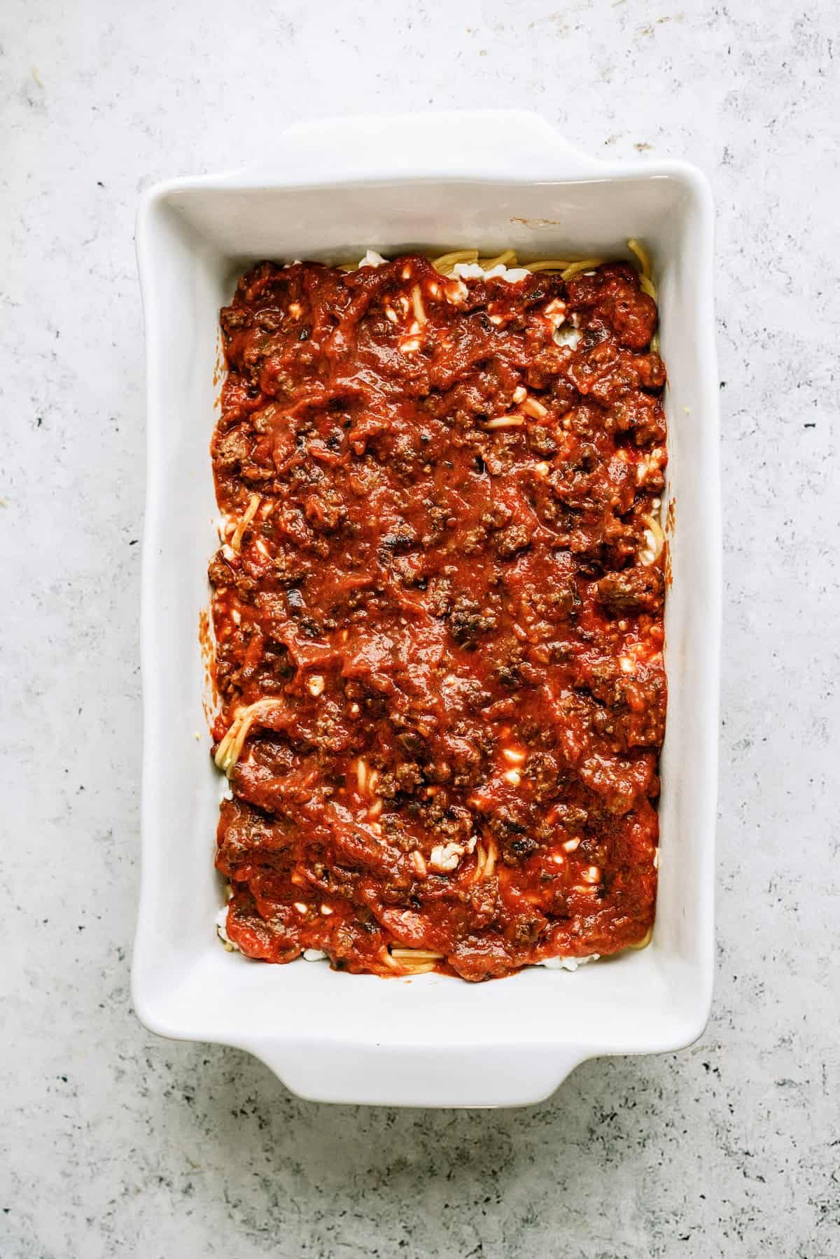 A rectangular white baking dish filled with lasagna topped with a layer of meat sauce, viewed from above on a light-colored surface.