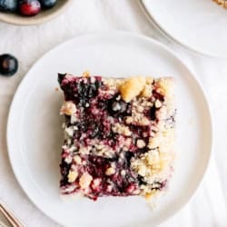 A square piece of blueberry crumb cake sits on a white plate, with fresh blueberries in a bowl nearby.