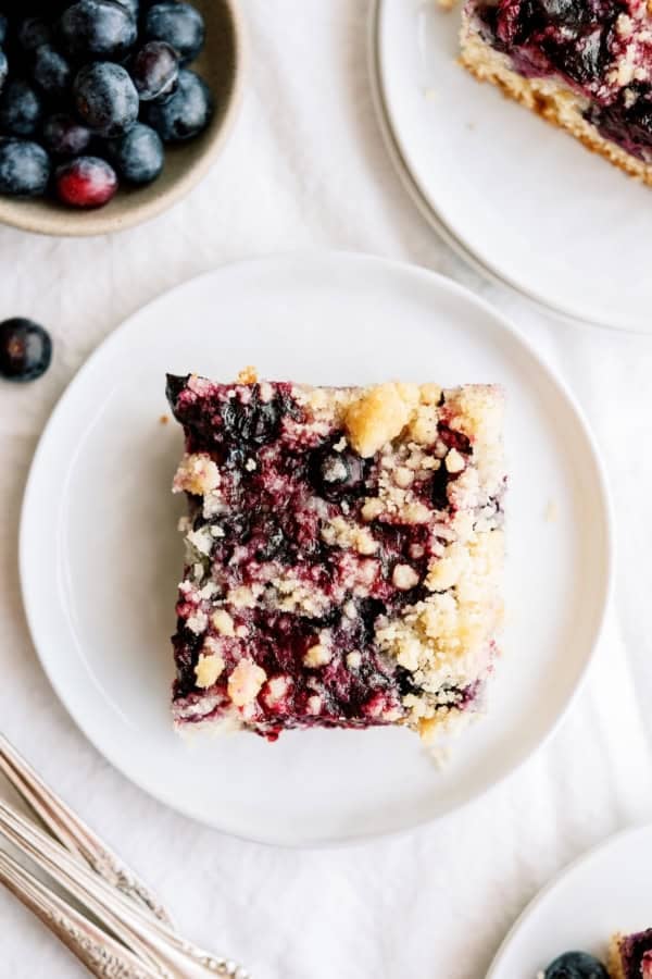 A square piece of blueberry crumb cake sits on a white plate, with fresh blueberries in a bowl nearby.