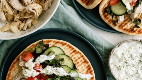 Two plates with flatbread topped with shredded chicken, cucumber, tomato, lettuce, and white sauce. A bowl of shredded chicken and a small bowl of sauce are nearby.