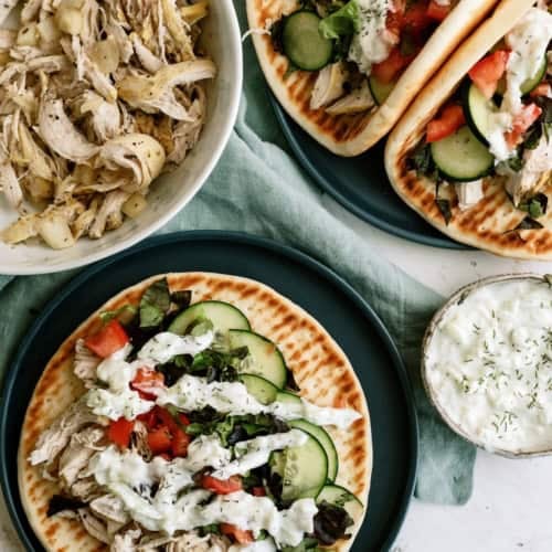 Two plates with flatbread topped with shredded chicken, cucumber, tomato, lettuce, and white sauce. A bowl of shredded chicken and a small bowl of sauce are nearby.