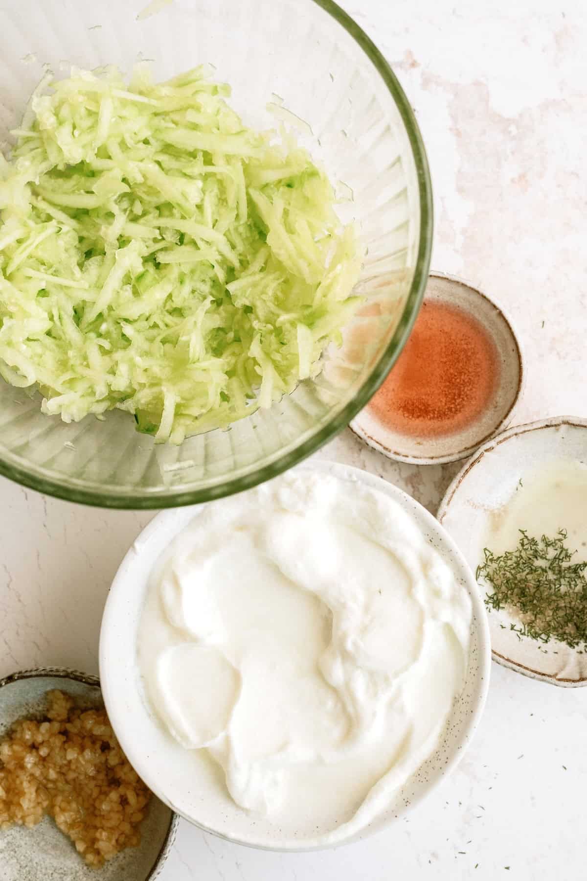 A bowl of grated cucumber, a bowl of plain yogurt, minced garlic, vinegar, salt, and dried dill on a white surface.