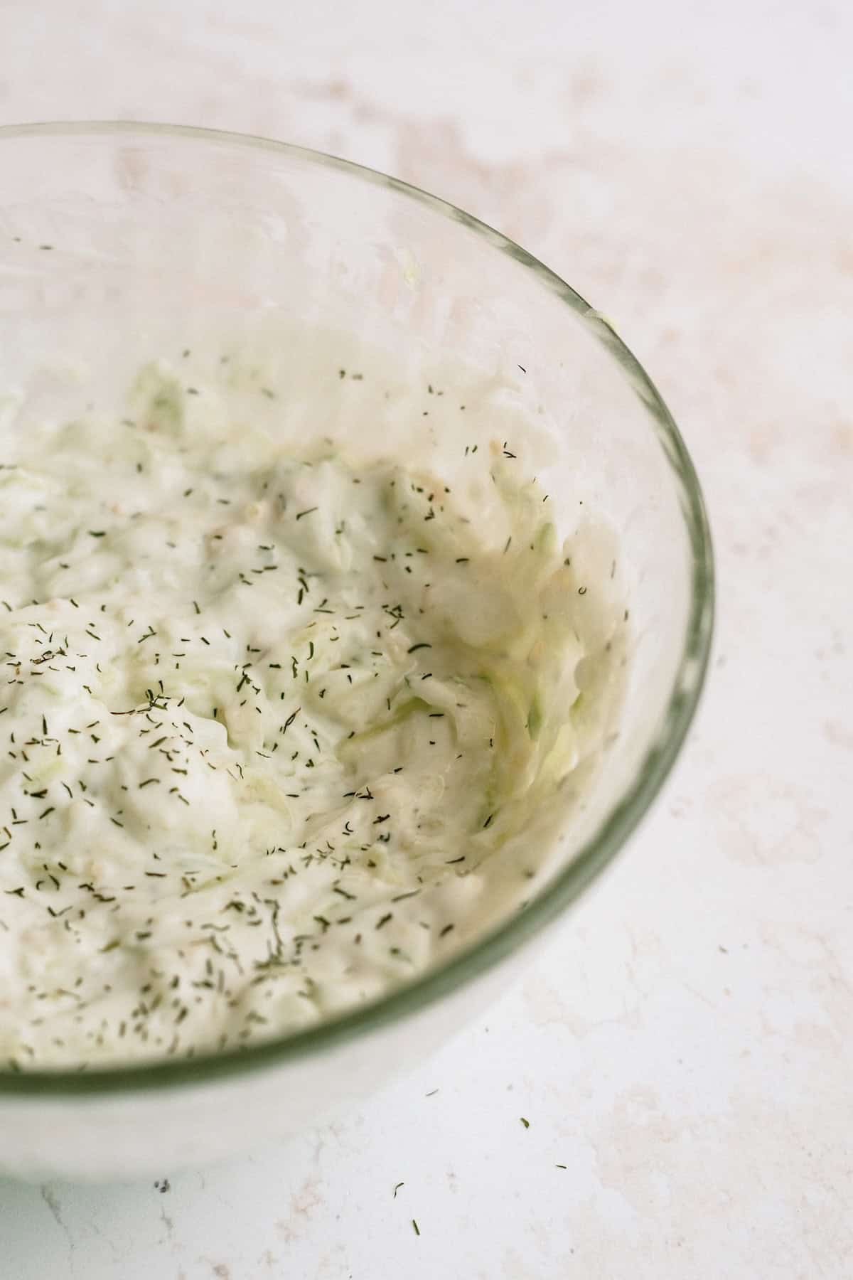 A clear glass bowl containing creamy cucumber salad garnished with dried dill, placed on a light-colored surface.
