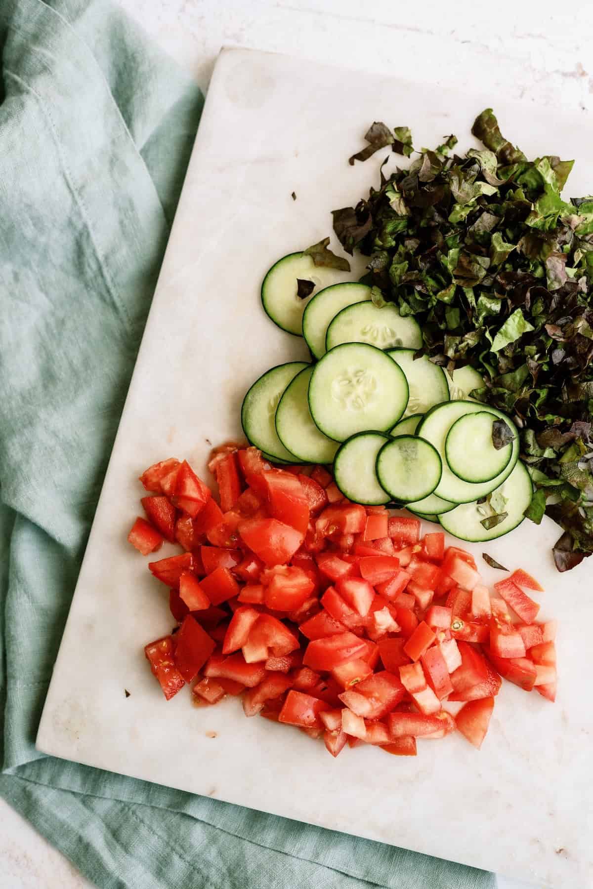 Chopped lettuce, sliced cucumbers, and diced tomatoes arranged on a white cutting board with a green cloth nearby.