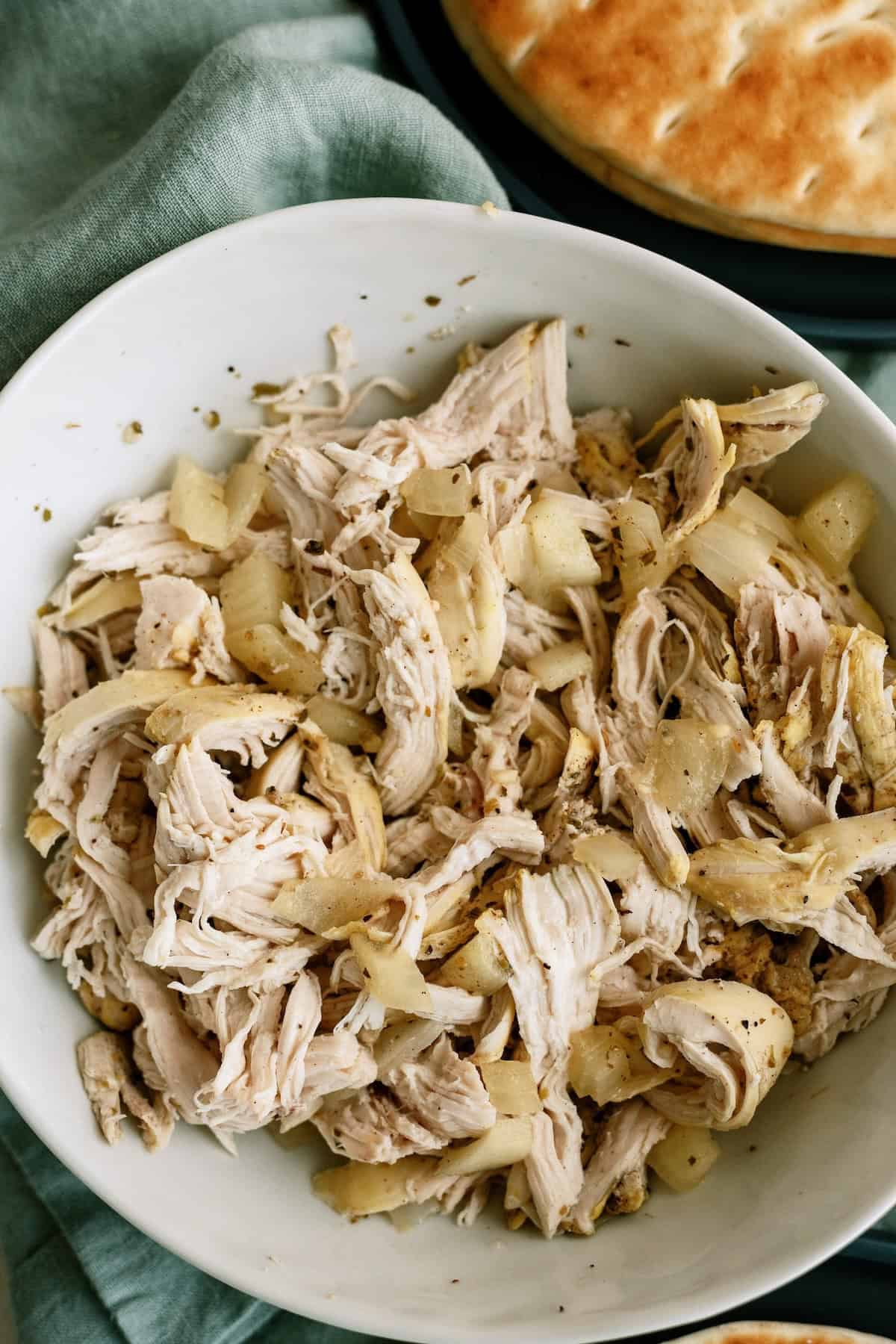 Shredded cooked chicken mixed with chopped onions in a white bowl, placed on a green cloth.